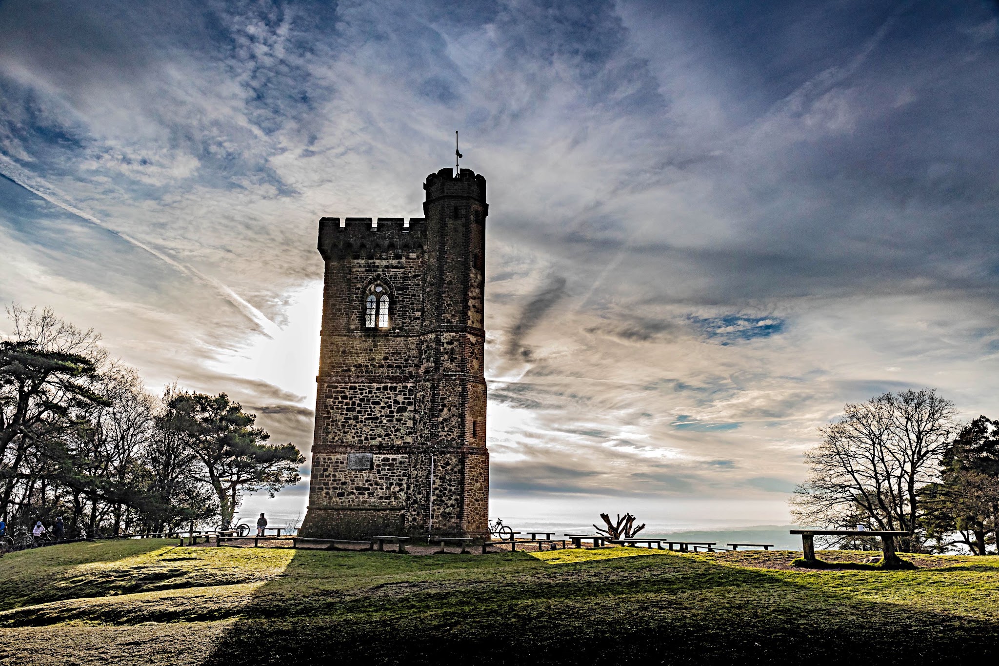 Cycling Leith Hill, England - Leith Hill tour at sunset with sun rays shining out from behind it, tall stone tower with small flag poll atop it, grass in front of tour and benches to side of it