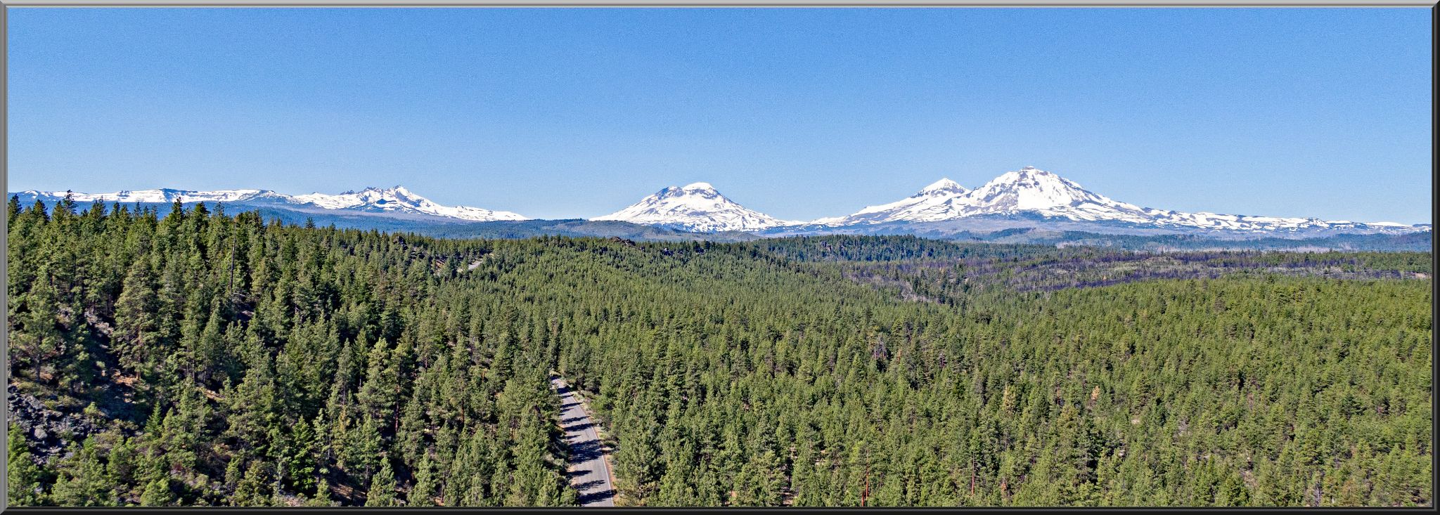aerial drone view of Three Creek Lake Road cut through the center of alpine forest, Three Sisters Volcanic Peaks in horizon