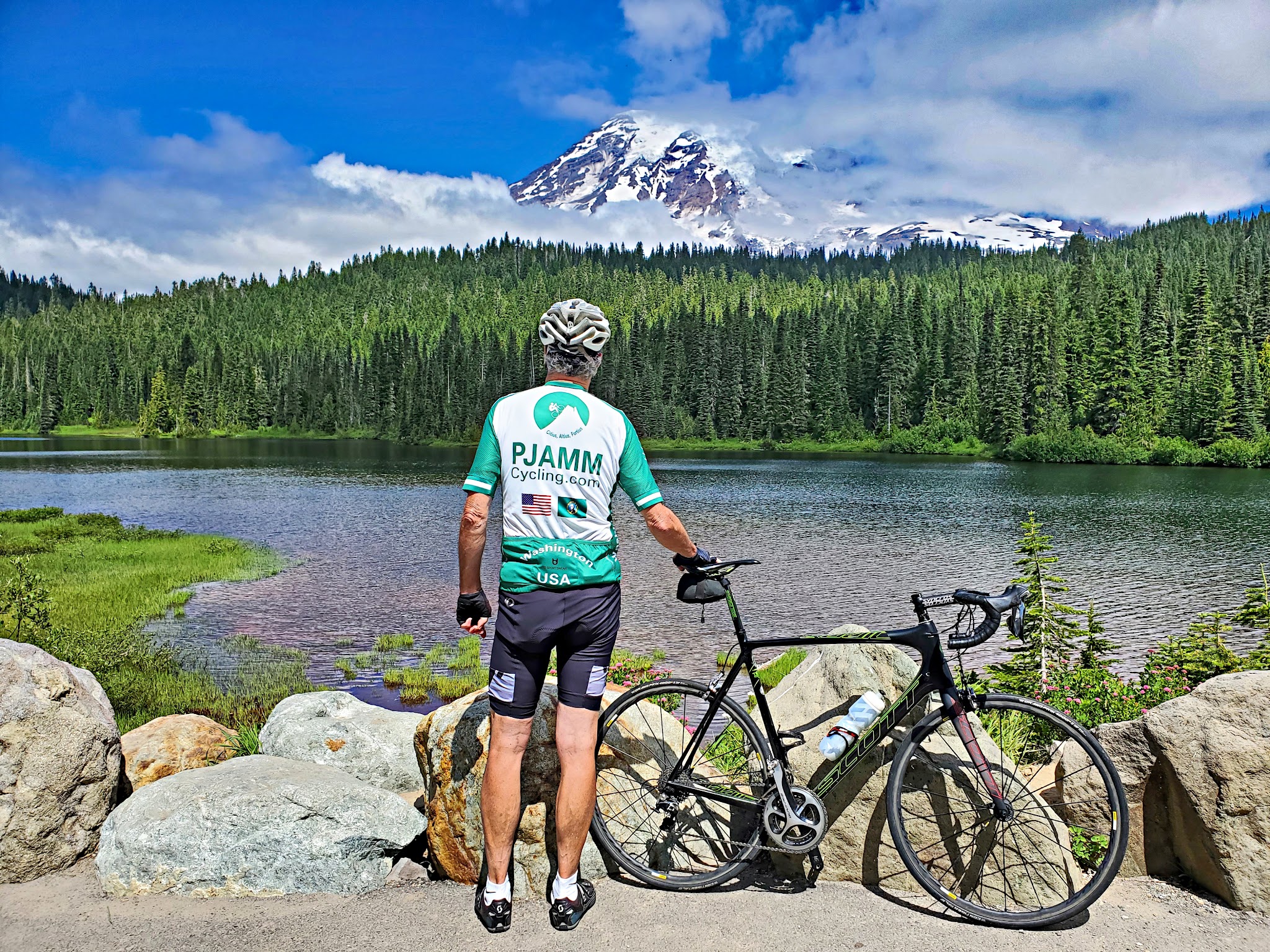 Cycling Mt. Rainier, Stevens Canyon, Washington - cyclist wearing PJAMM Cycling jersey standing with bike facing Reflection Lakes, evergreen trees and snow capped mountain behind clouds in distance across the lake