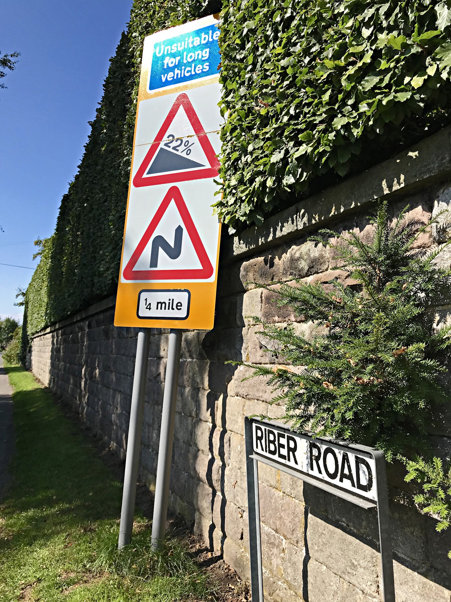 Cycling Riber, England - road sign for Riber Road against a brick wall partially covered in greenery, next to that a second road sign stating "Unsuitable for long vehicles, 22% grade, windy road for 1/4 mile"