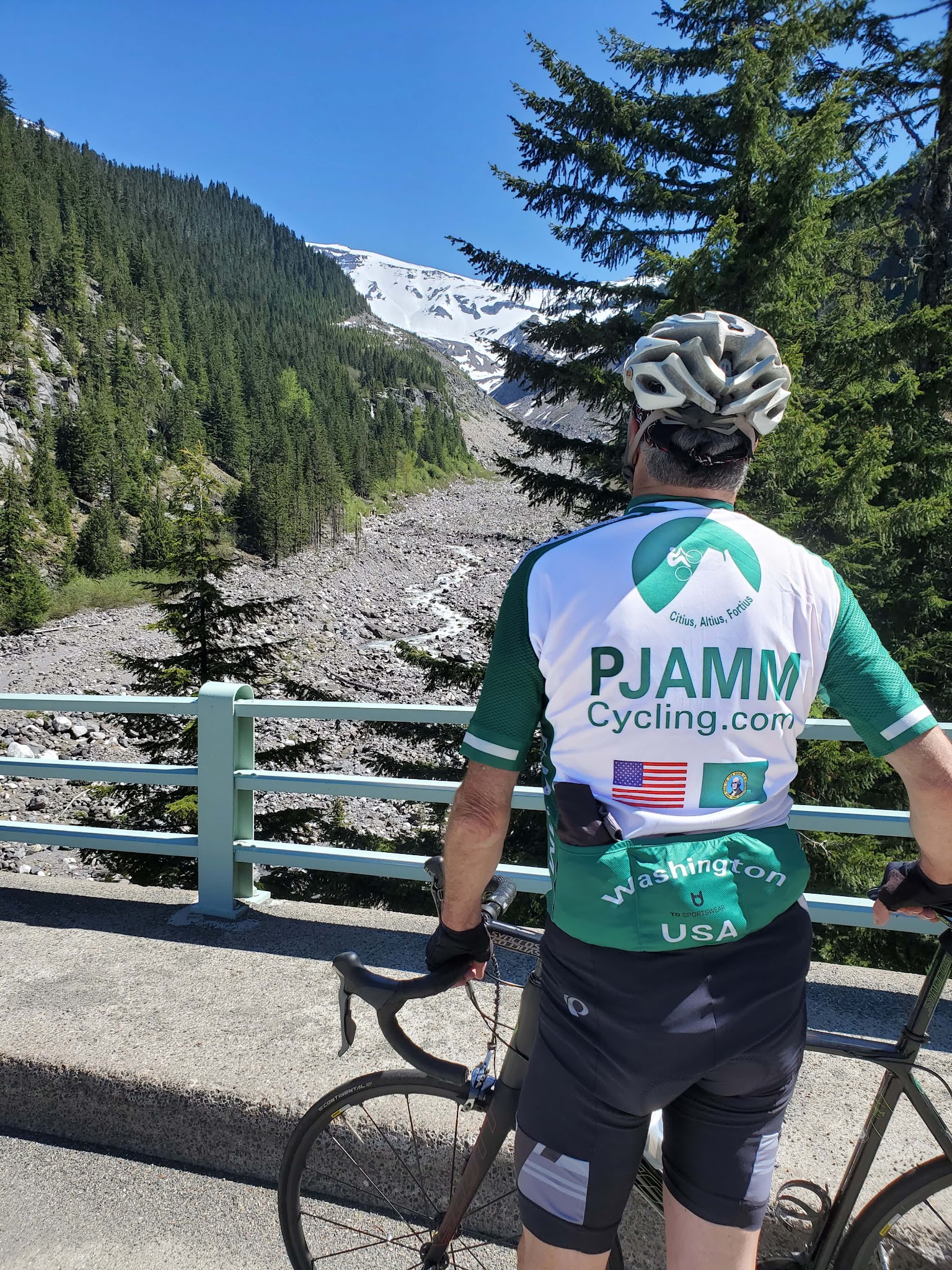 Cycling Mt. Rainier from Nisqually River Entrance - cyclist wearing white and green PJAMM Cycling jersey stands with bike looking out at creek bed from the Nisqually River Bridge