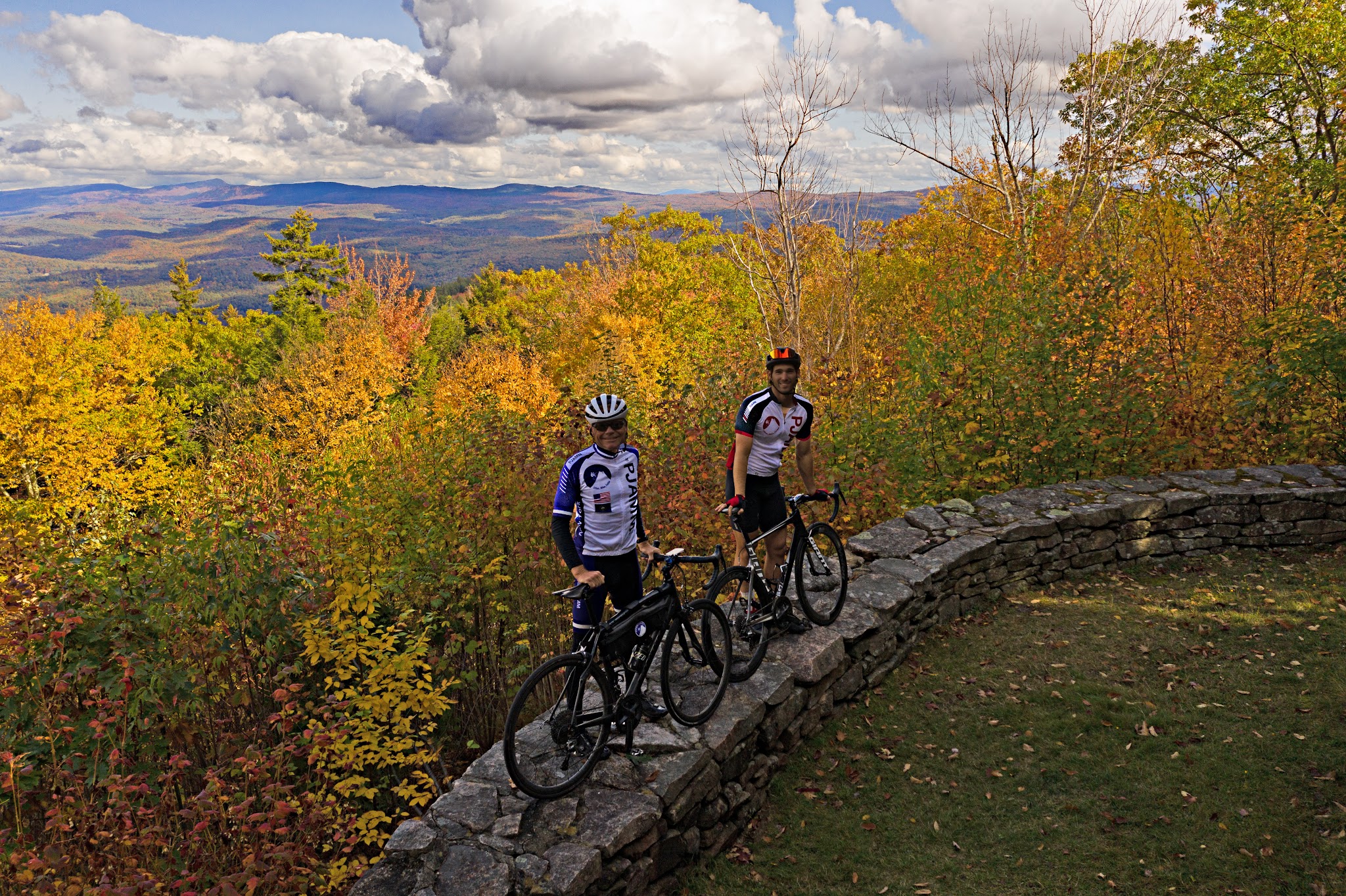 Cycling Mt. Ascutney, Vermont - two PJAMM Cyclists stand on low stone retaining wall with their bikes surrounded by bright fall colors