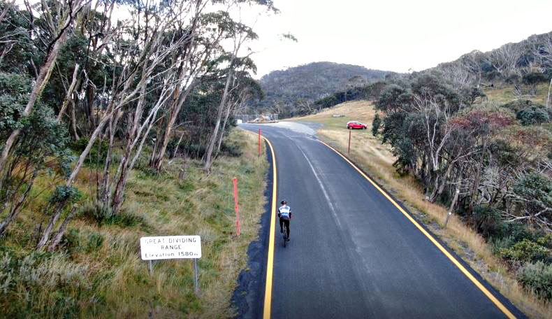 aerial drone view shows PJAMM Cyclist riding on eucalyptus-lined roadway next to sign for Great Dividing Range