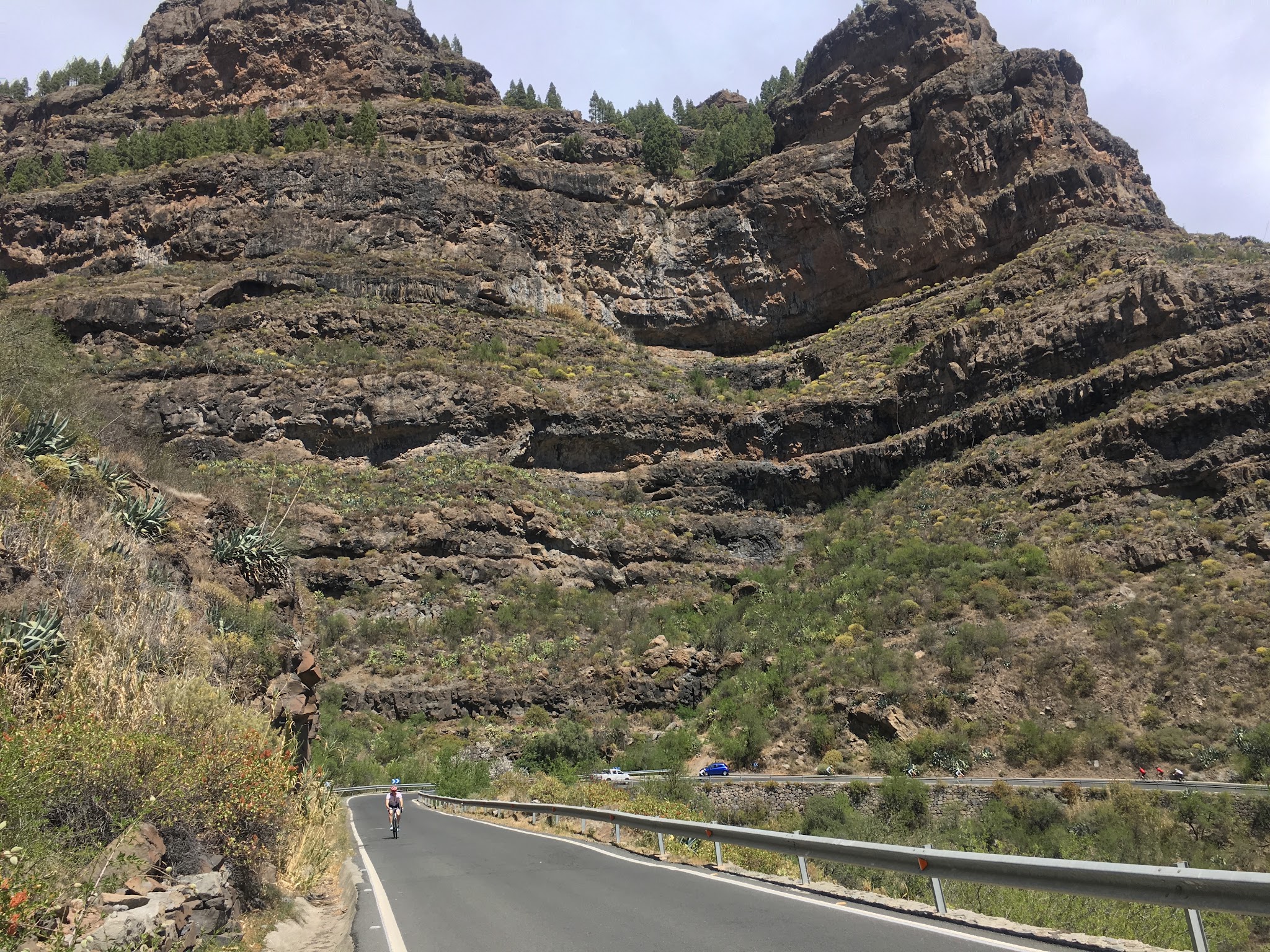PJAMM cyclist riding up roadway with amazing rock formations in mountainside behind him
