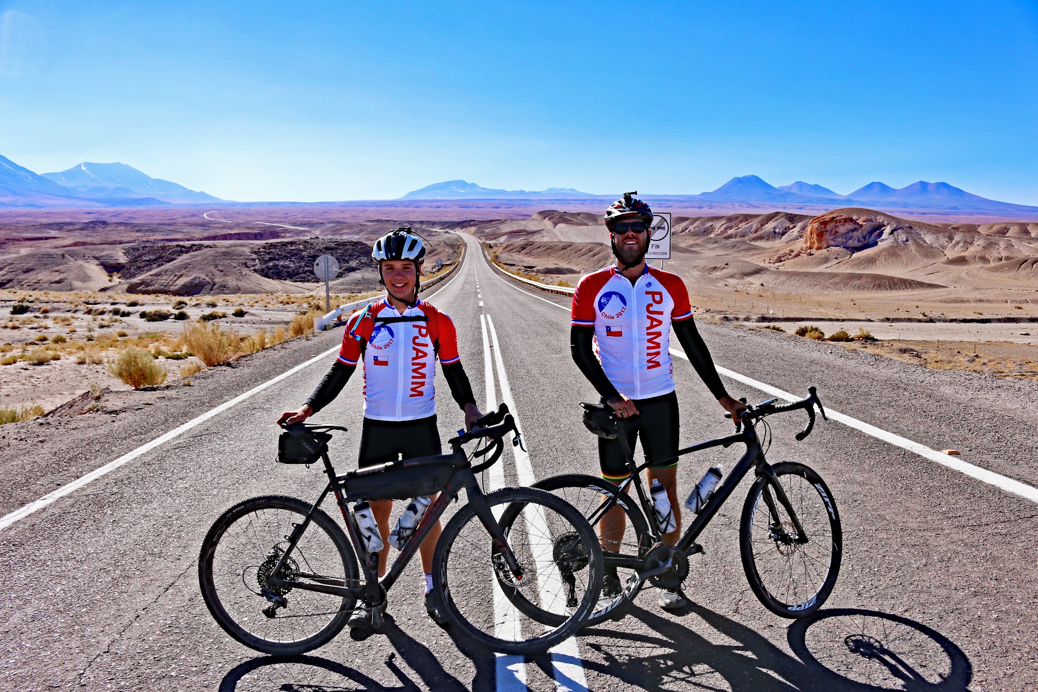Atacama - two PJAMM Cyclists stand with bikes in the middle of straight stretch of highway roadway in the Atacama Desert