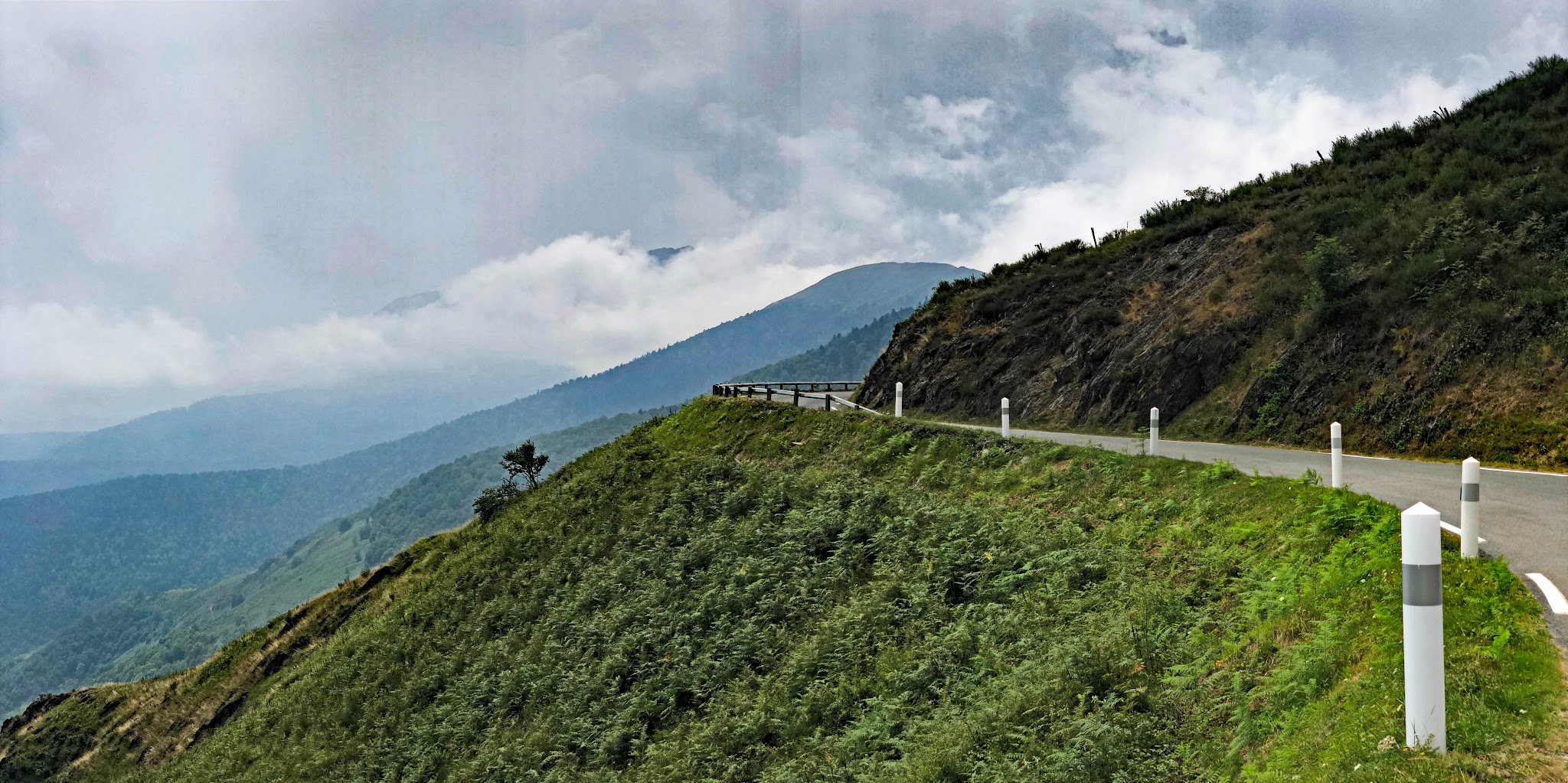 panoramic view of one-lane mountain road, lush green landscape on mountainside, tall mountains in background, bright white clouds