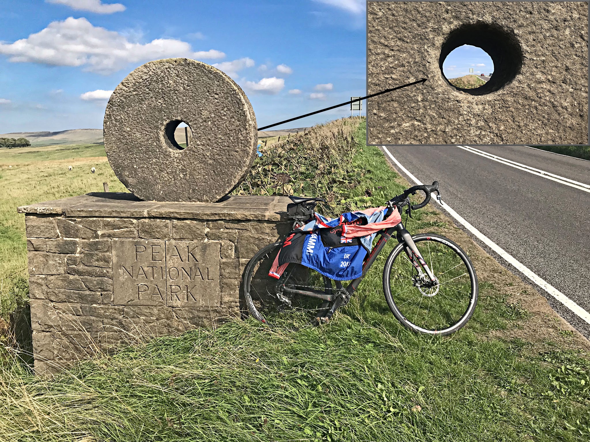 Cycling Cat & Fiddle, England - stone circular monument along roadway with bike parked in front of it with PJAMM Cycling jersey draped over it, monument says "Peak National Park" and has a hole in the center through which you can see the peak in the distance