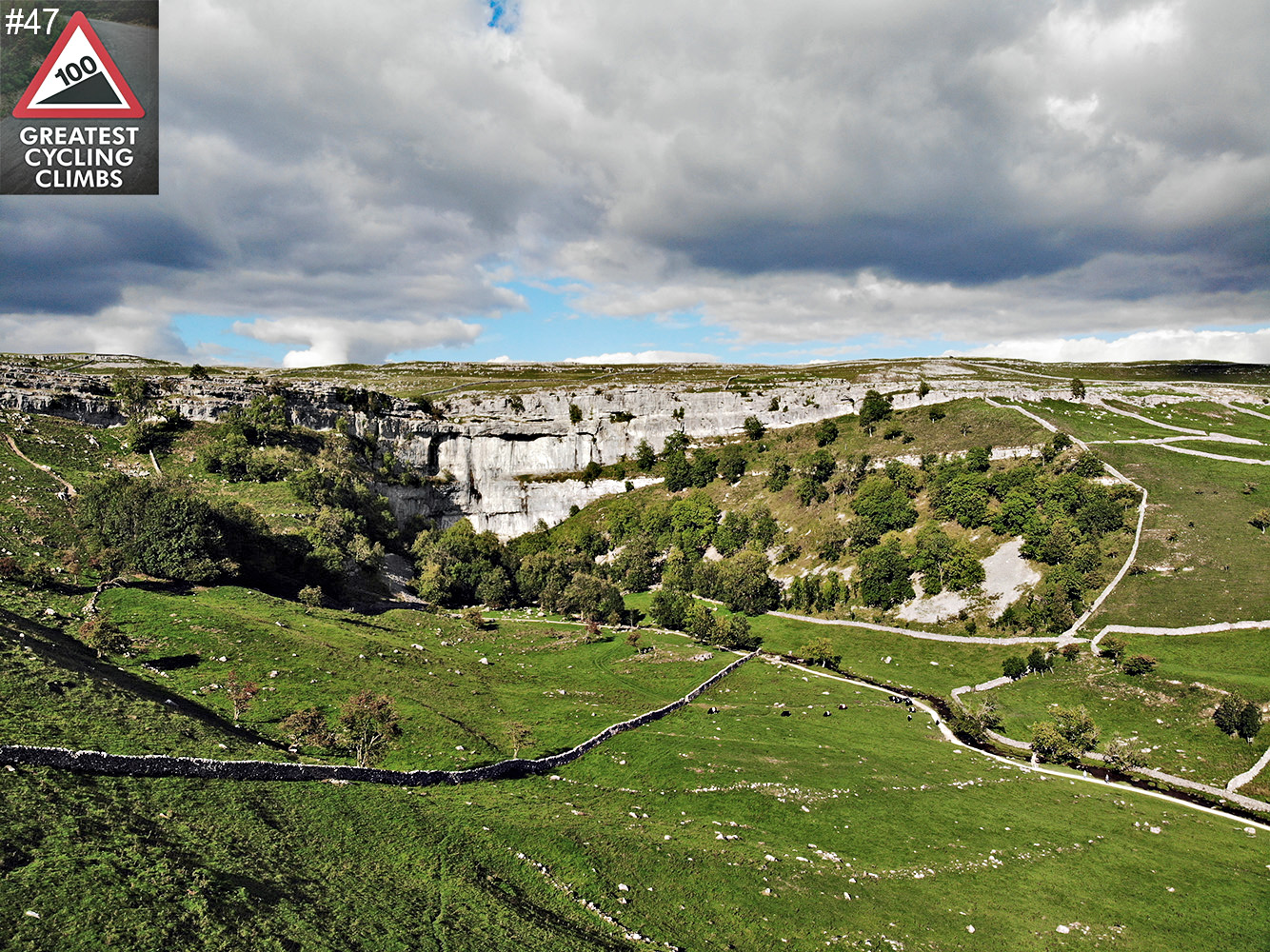Cycling Malham Cove - rolling green hills leading to large limestone rock wall, cloudy blue sky above; Simon Warren's 100 Greatest Cycling Climbs #47 logo in corner