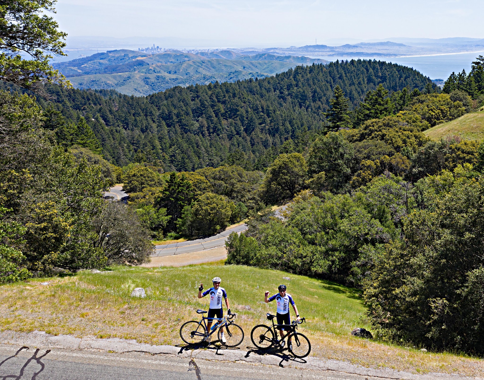 Cycling Mt. Tamalpais from Panoramic Highway South Cycling Mt. Tamalpais from Panoramic Highway South - aerial drone view of two cyclists wearing PJAMM Cycling jerseys and holding their bikes, waving at camera, standing along roadside overlooking rolling hills covered in forestation and ocean beyond