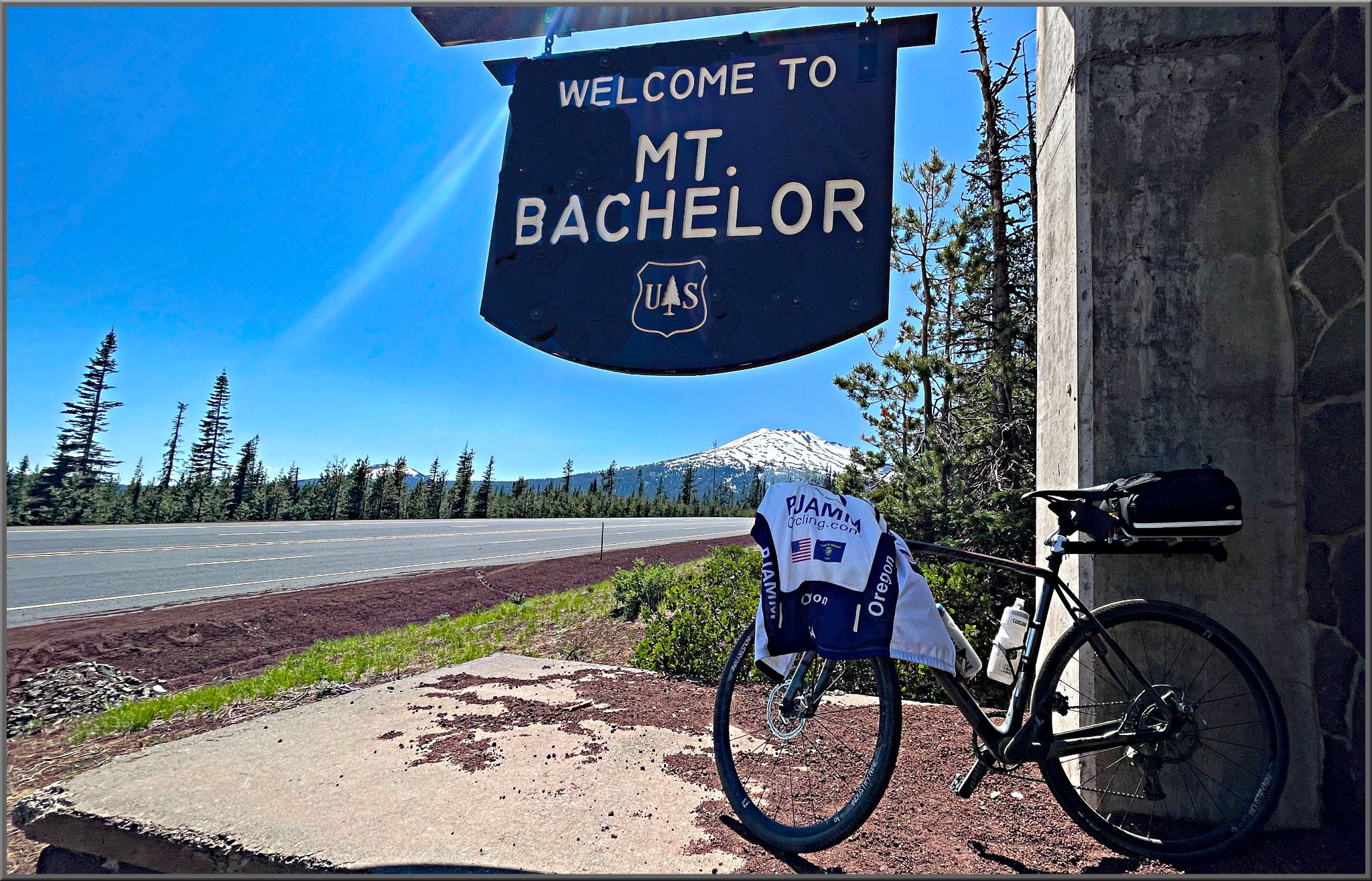 Bike with PJAMM Cycling Jersey draped over it parked underneath US Forest Service sign reading "Welcome to Mt. Bachelor"