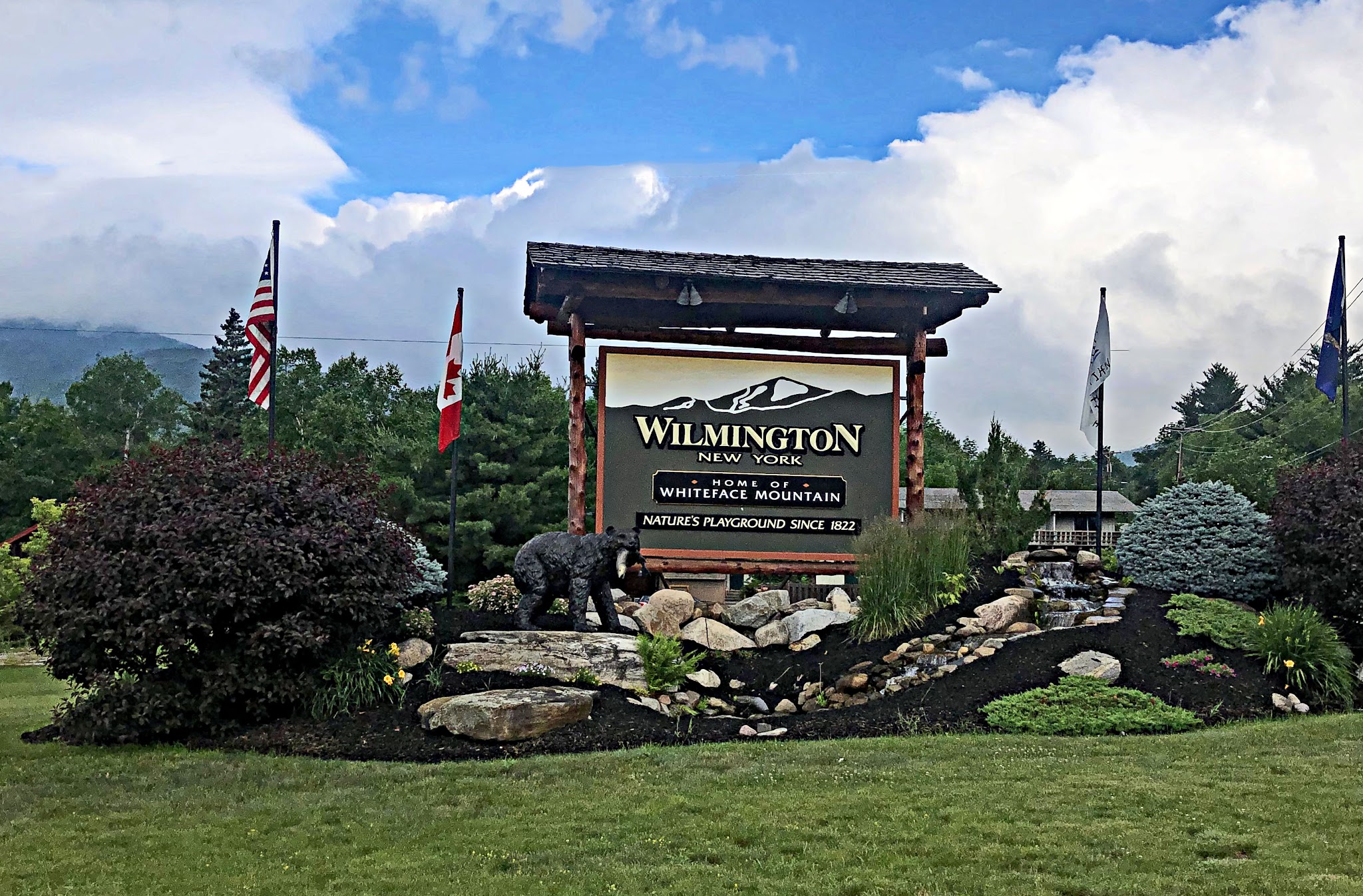 Cycling Whiteface Mountain - large decorative Wilmington sign surrounded by ornate landscaping, flags, and a bear statue 