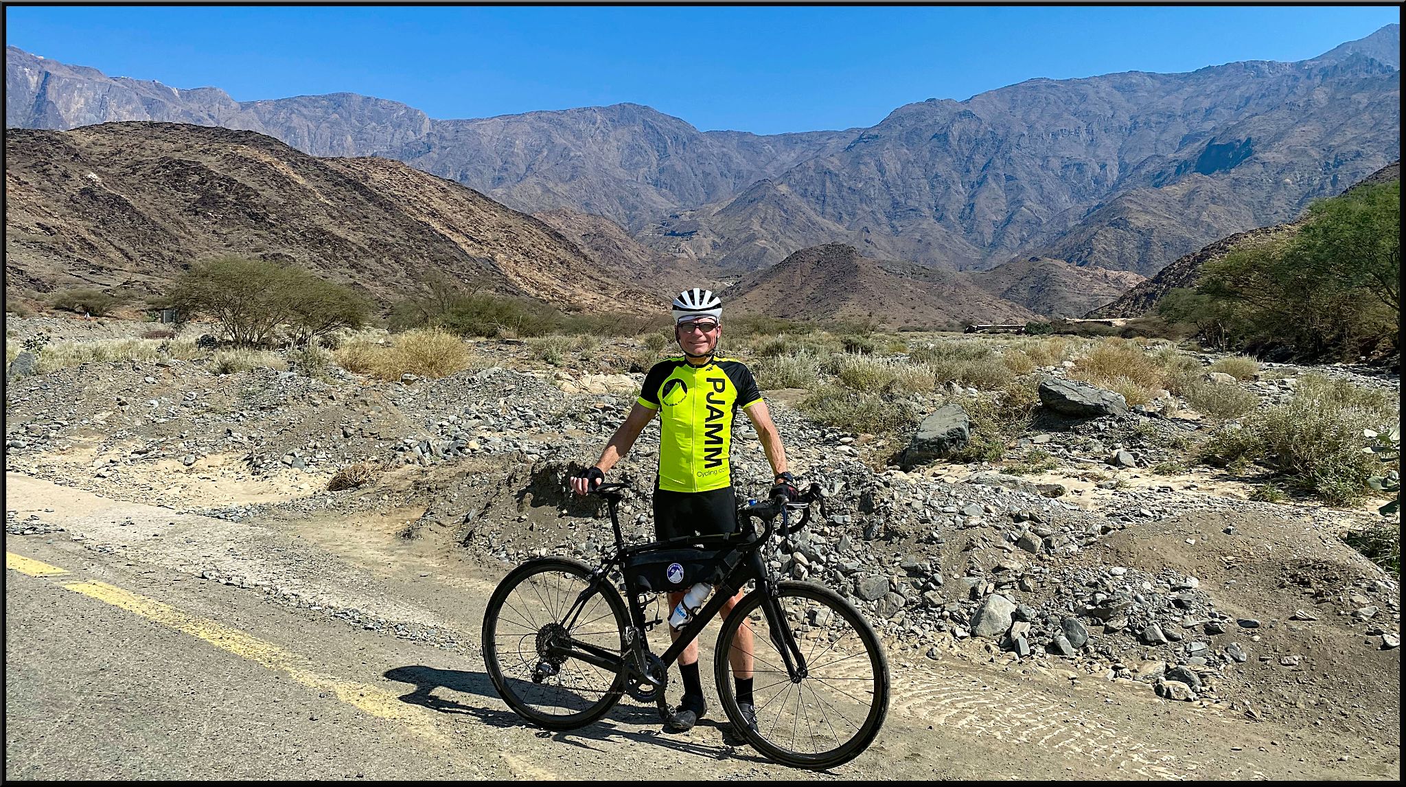 Cycling Beth-rah Pass, Saudi Arabia - PJAMM Cyclist John Johnson stands with bike in front of desert landscape, Asir mountains behind