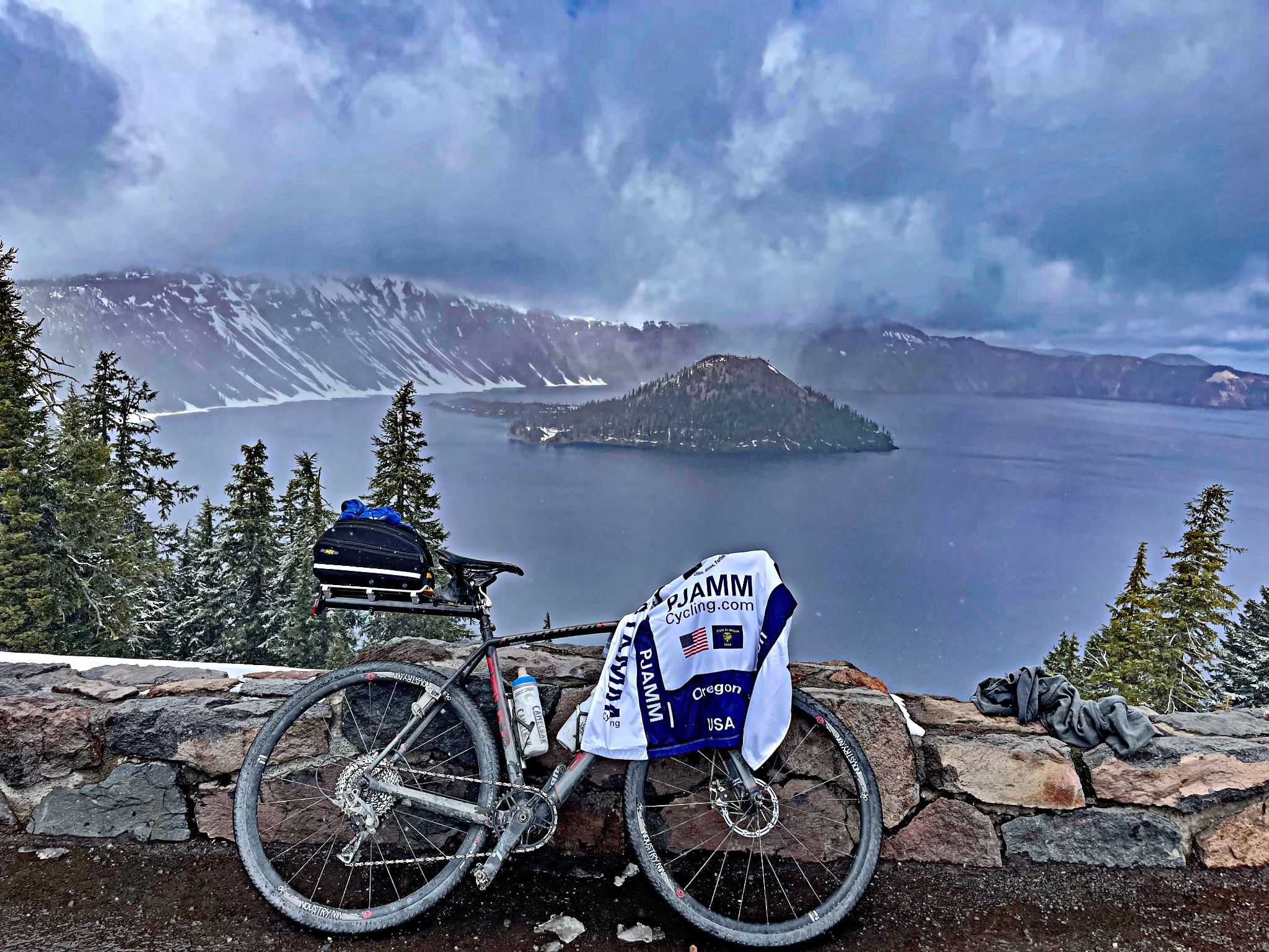 Bike with PJAMM Cycling jersey draped over it parked against low stone retaining wall overlooking Crater Lake and the island in the center, Oregon Top Bike Climb