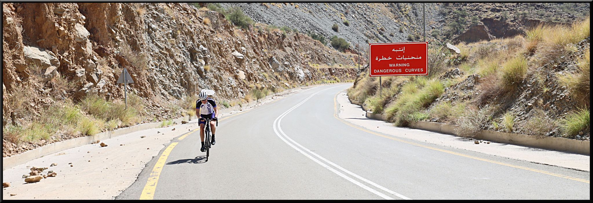 Cycling Bani Horairah - PJAMM Cyclist rides on flat portion of two-lane highway roadway, surrounded by rocky walls