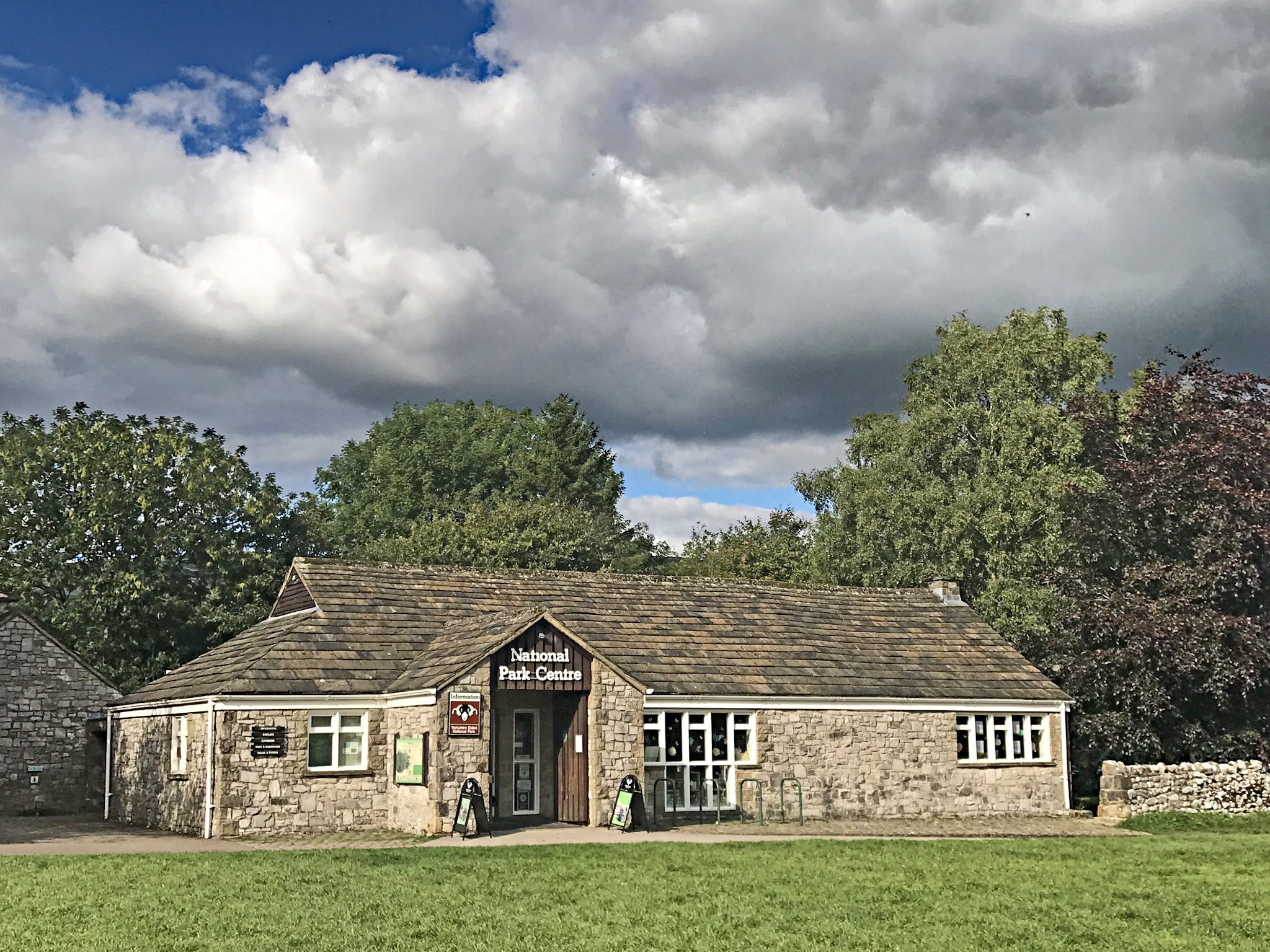 Cycling Malham Cove - stone building with sign "National Park Center", green grass, large green trees behind building, blue sky with large white clouds