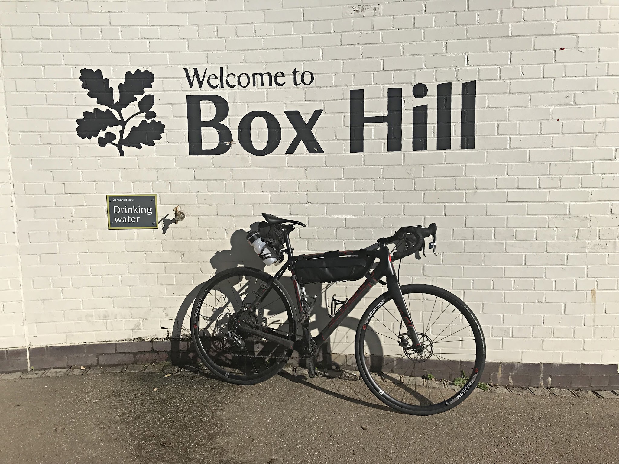 Cycling Box Hill - white brick wall with "Welcome To Box Hill" painted on it, road bike leaning against wall next to drinking faucet