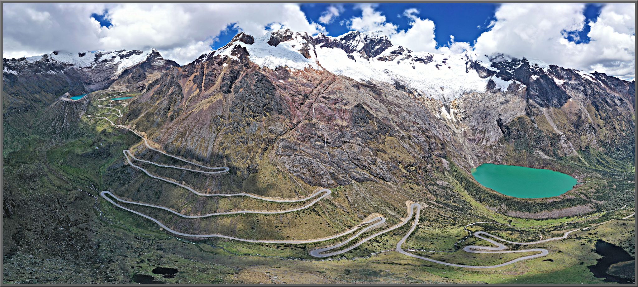 Cycling Punta Olimpica East, Peru - panoramic view shows the roadway snaking up the mountainside, bright green glacial lake on right of picture below snow-capped mountain peaks