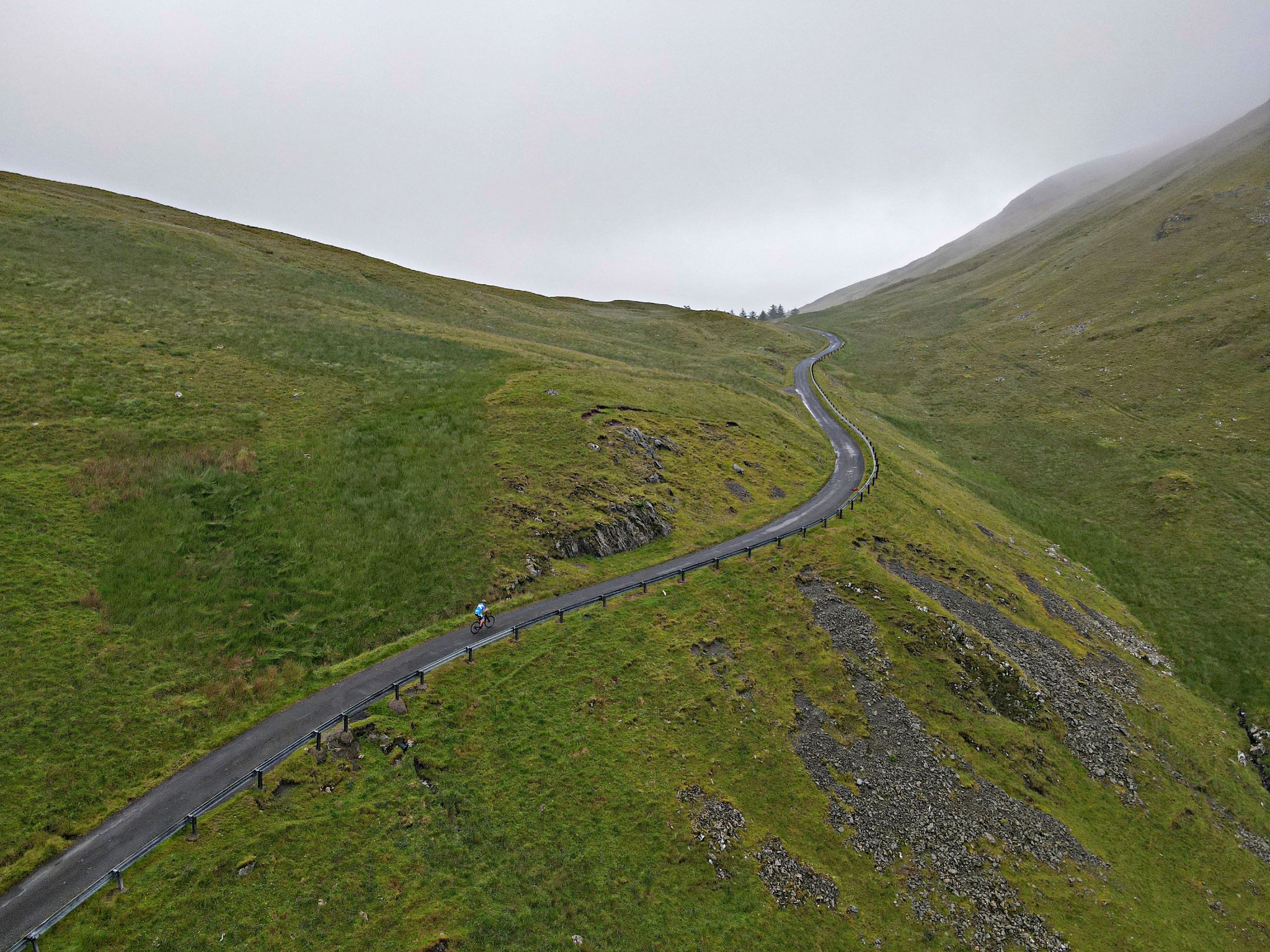 aerial drone view of PJAMM Cyclist climbing alone up the roadway, snaking through the green Scottish hillsides